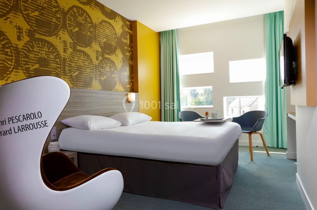 Chambre d'hôtel lumineuse avec un grand lit, fauteuil design, bureau, et fenêtres laissant entrer la lumière naturelle.