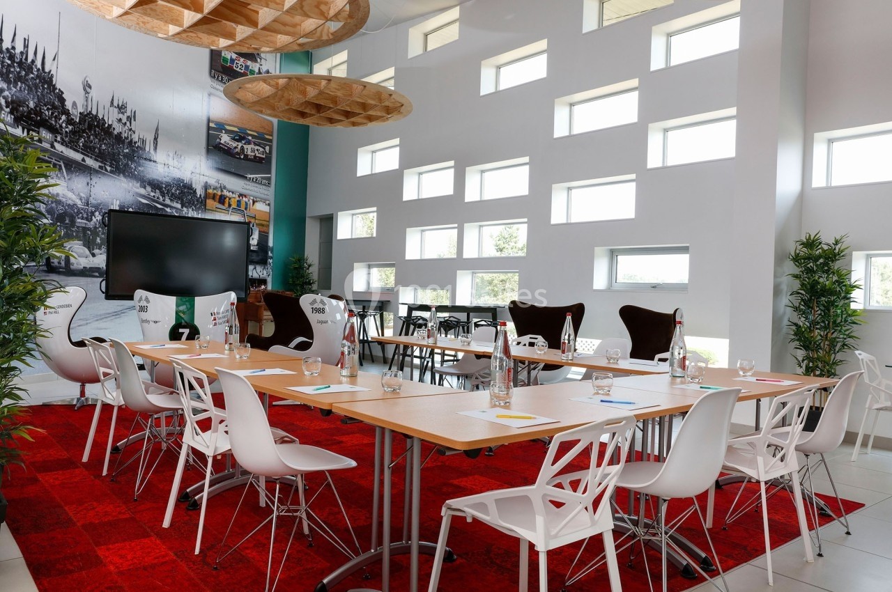 Salle de réunion lumineuse avec tables en bois, chaises blanches, écran, plantes et décoration murale.