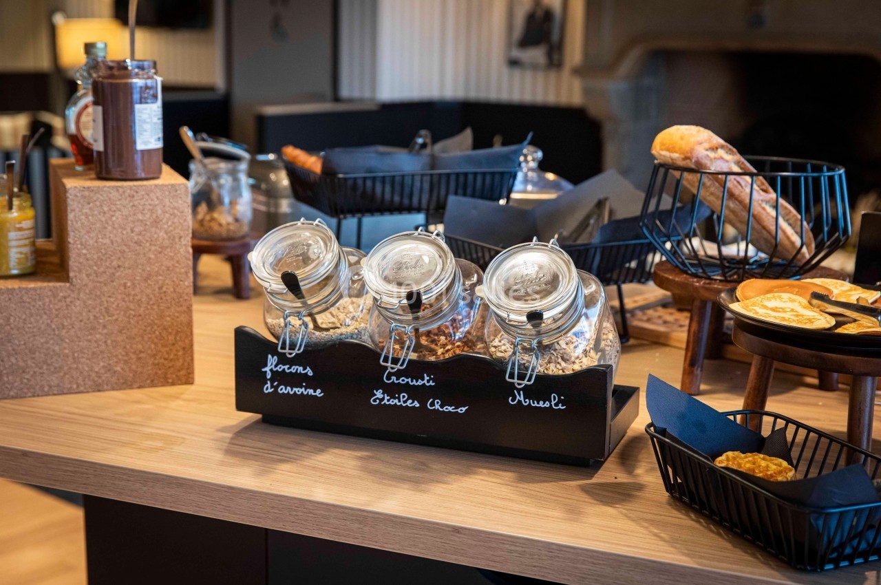 Pots transparents contenant des céréales, disposés sur un comptoir avec des pains et condiments en arrière-plan.