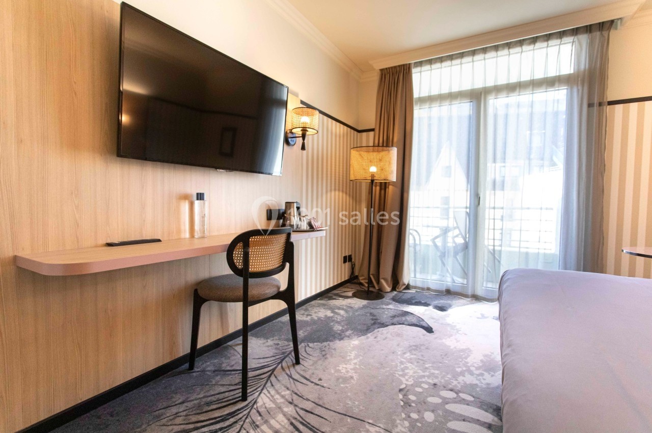 Chambre d'hôtel lumineuse avec bureau, chaise, télévision murale, lit et balcon donnant sur l'extérieur.