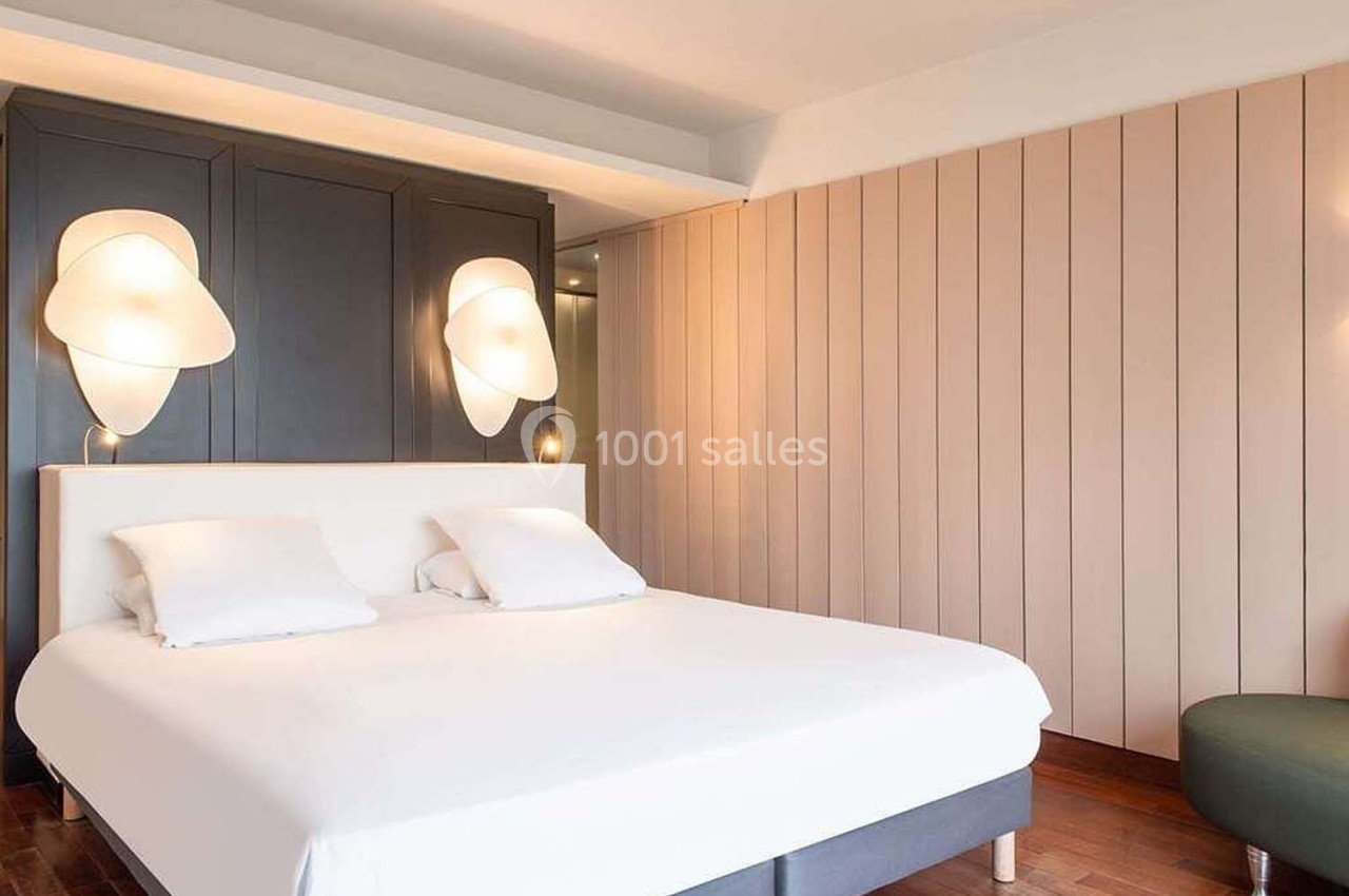 Lit double avec draps blancs dans une chambre moderne, éclairée par des appliques murales et décorée de panneaux en bois…