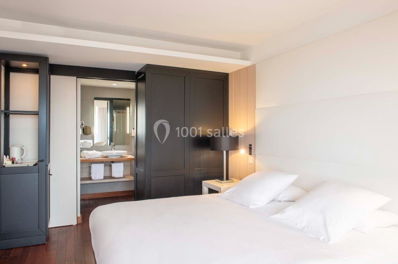 Chambre lumineuse avec lit double, armoire noire, table de chevet et vue sur une salle de bain moderne en arrière-plan.