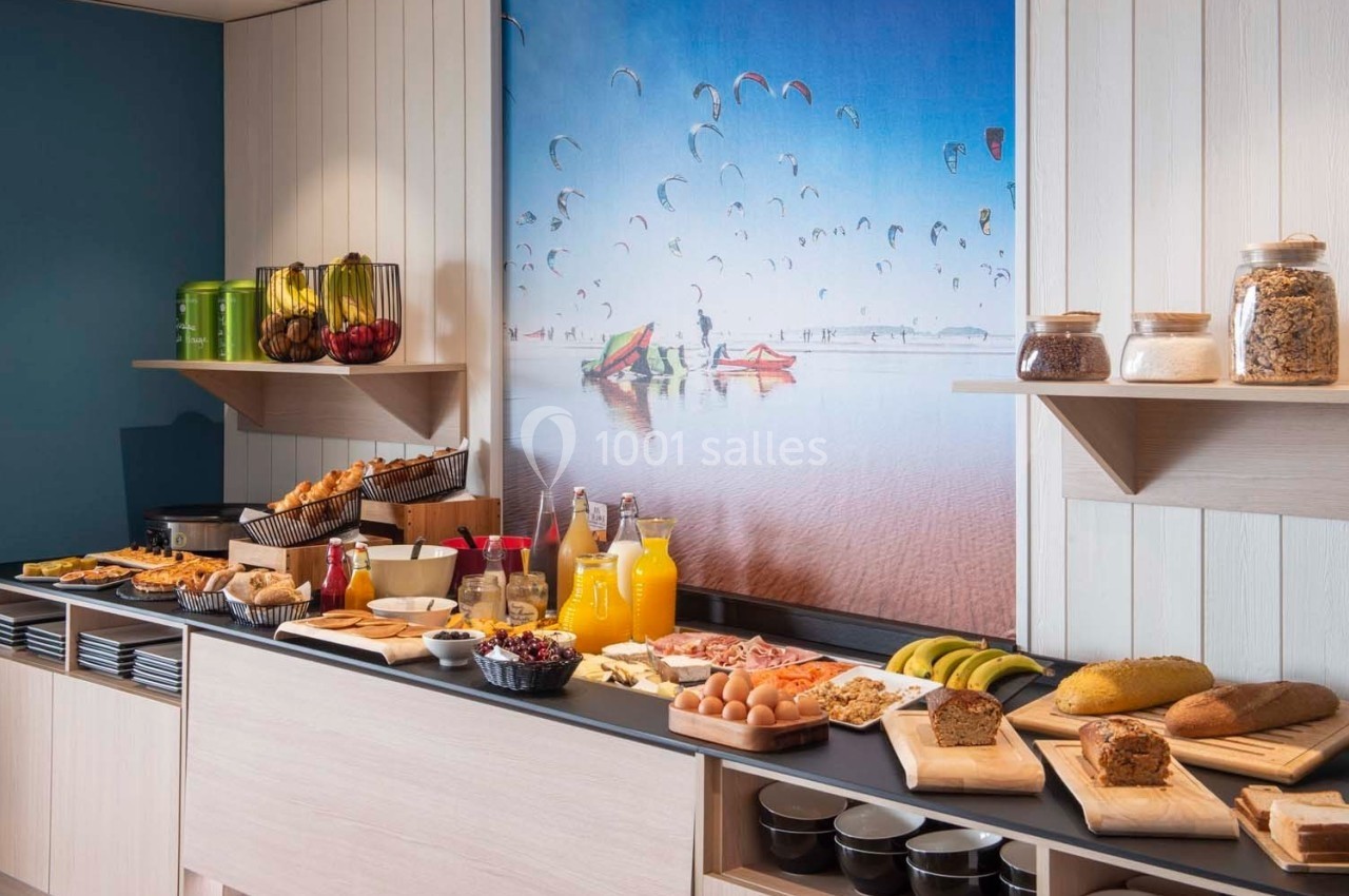 Buffet de petit-déjeuner avec viennoiseries, fruits, jus, céréales et pain, devant une photo de plage avec des cerfs-volants.