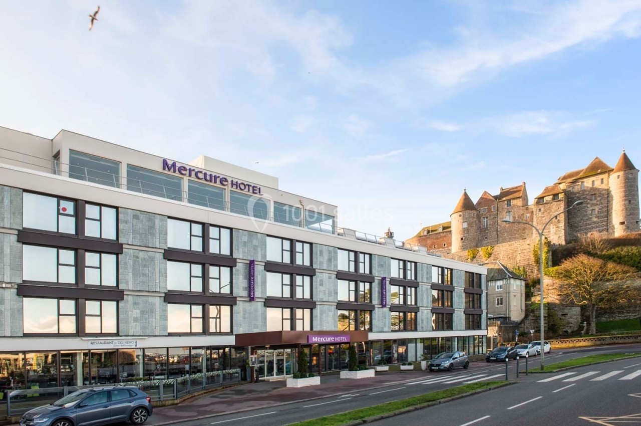 Façade d'un hôtel moderne avec un château en arrière-plan, situé près d'une route et sous un ciel dégagé.