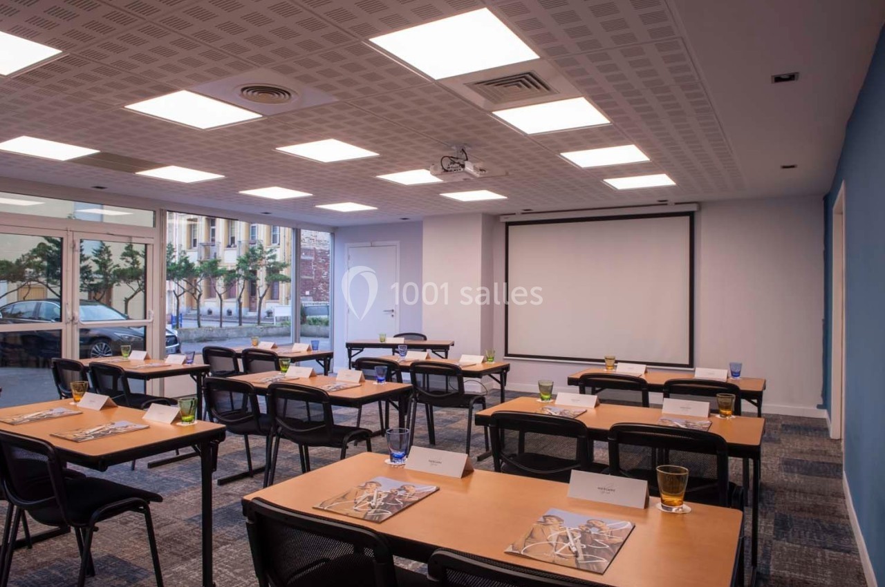 Salle de réunion lumineuse avec tables disposées en rangées, chaises noires, écran de projection et grandes fenêtres.