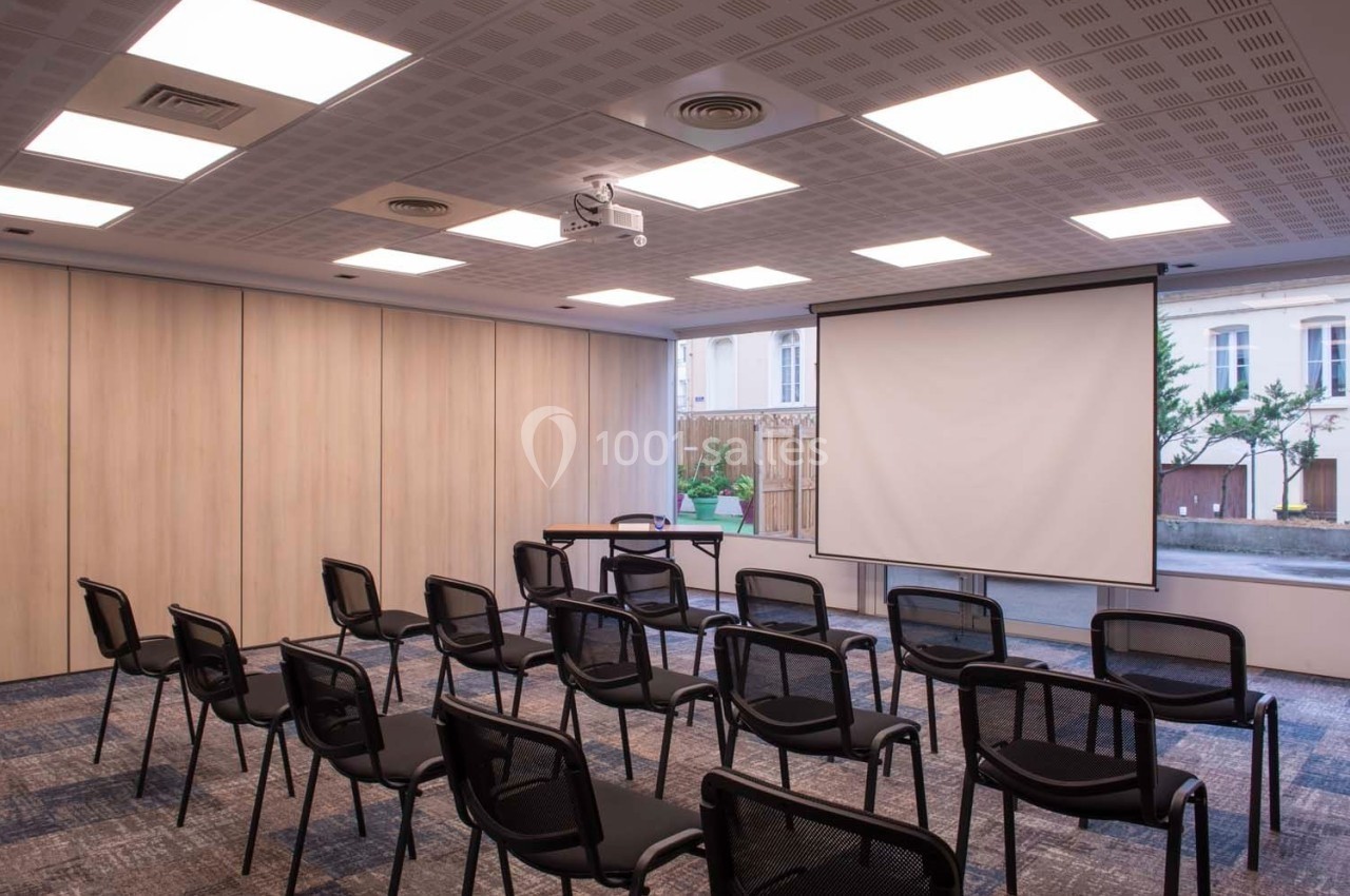Salle de réunion avec chaises alignées, écran de projection déployé et lumière naturelle entrant par une baie vitrée.