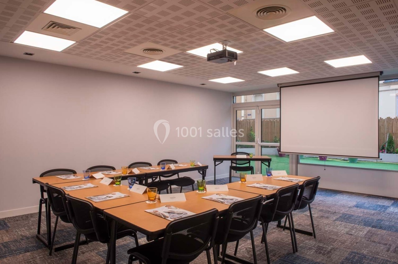 Salle de réunion lumineuse avec tables disposées en U, chaises noires, écran de projection et fenêtres donnant sur un espace…