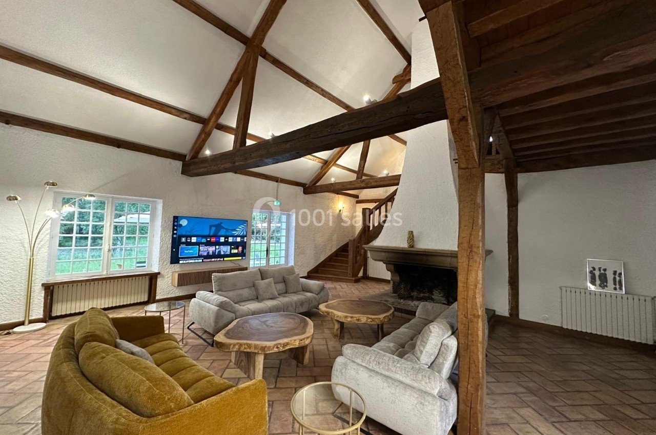 Salon spacieux avec poutres apparentes, cheminée, canapés et tables basses en bois, éclairé par des fenêtres.