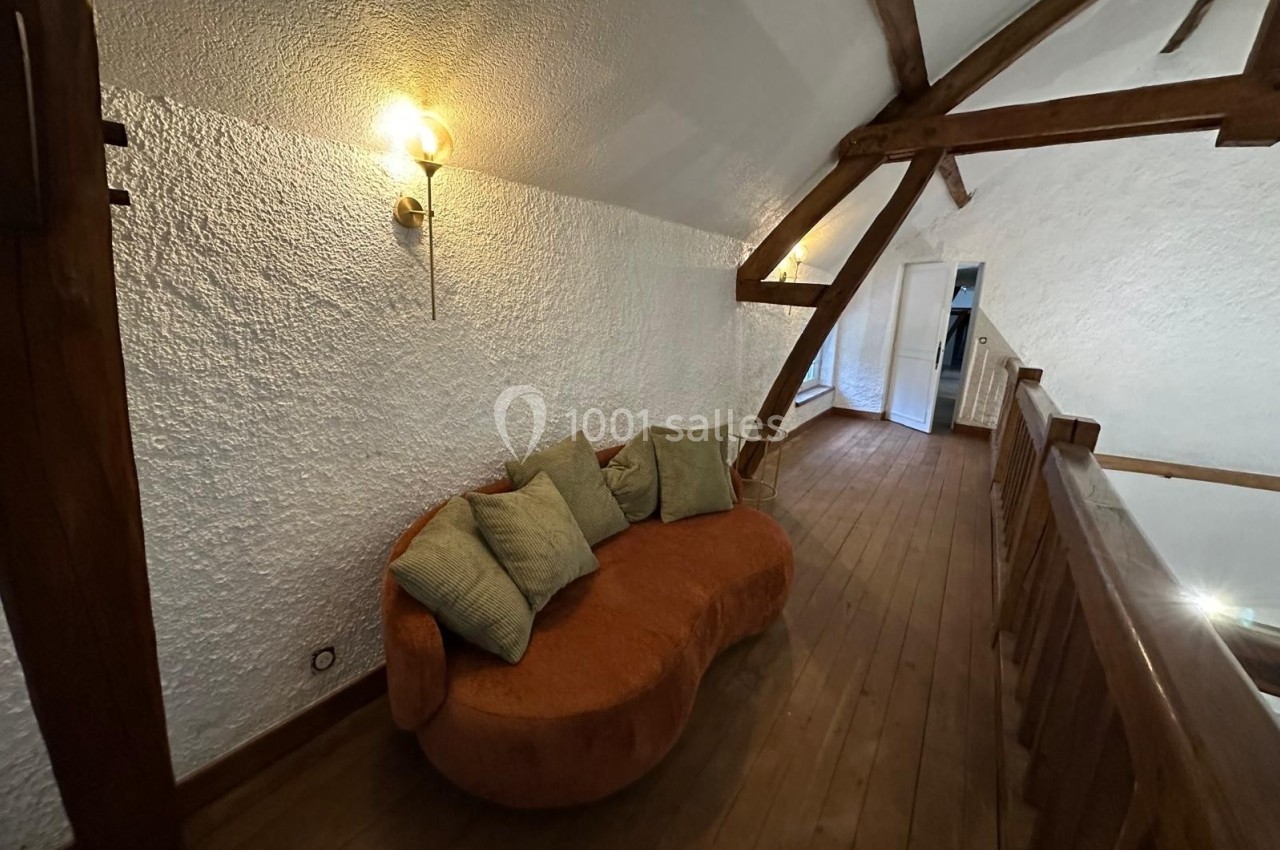Canapé orange avec coussins verts placé sur un parquet en bois, situé dans un espace mansardé avec poutres apparentes.