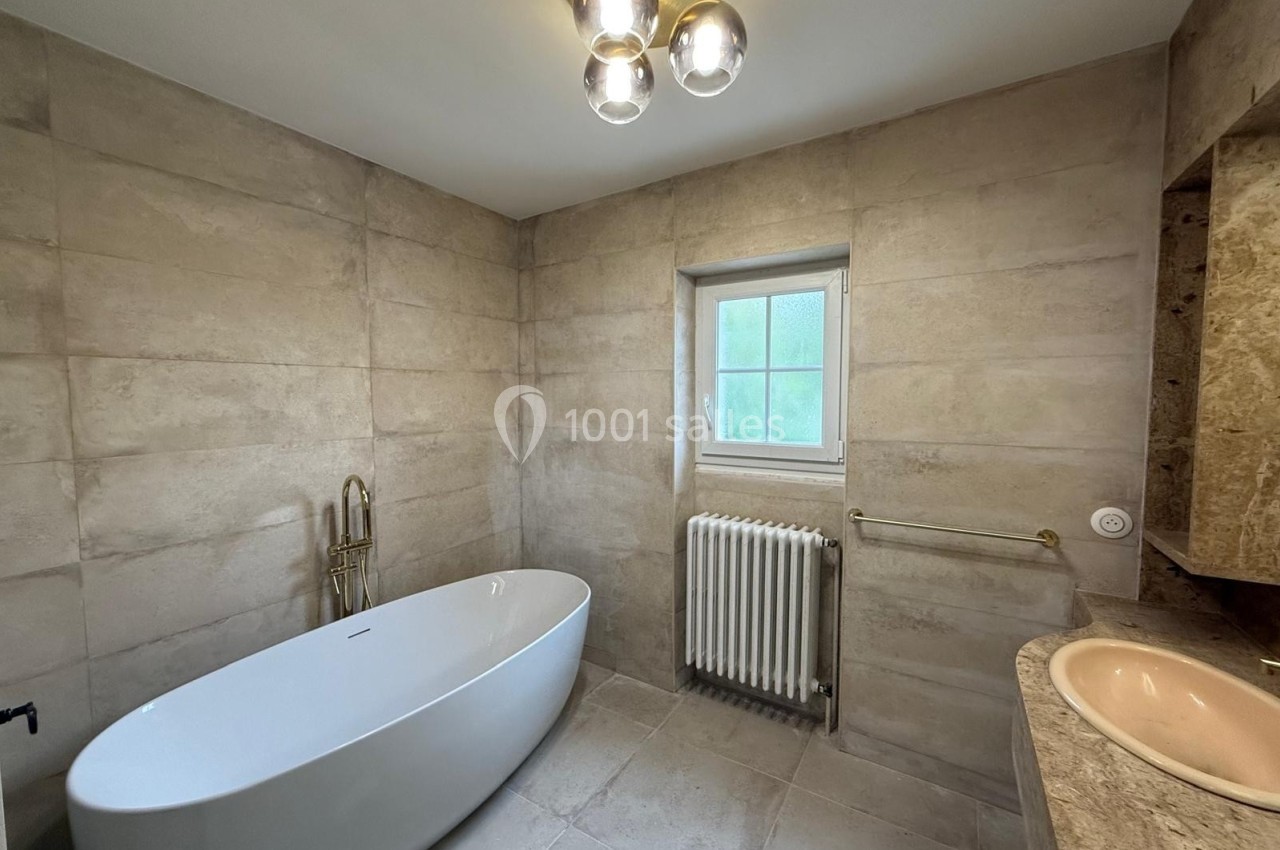 Salle de bain moderne avec baignoire îlot, double vasque, murs en carrelage beige et fenêtre laissant entrer la lumière…