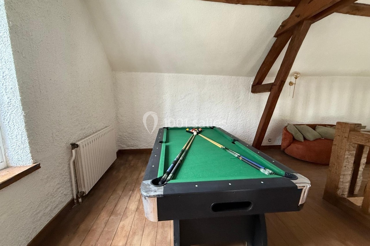 Table de billard avec tapis vert et deux queues posées dessus, située dans une pièce mansardée avec poutres apparentes.