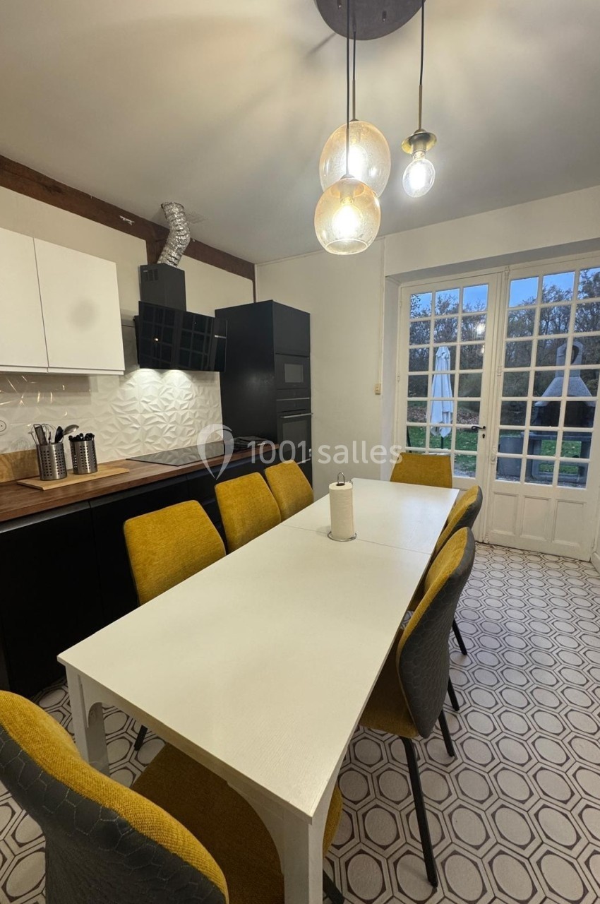 Salle à manger avec table blanche, chaises jaunes, cuisine moderne en arrière-plan et porte-fenêtre donnant sur un jardin.