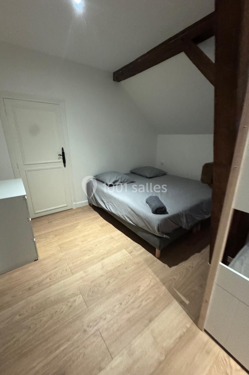 Chambre avec lit double, parquet clair, poutres apparentes et mobilier minimaliste dans un espace mansardé.