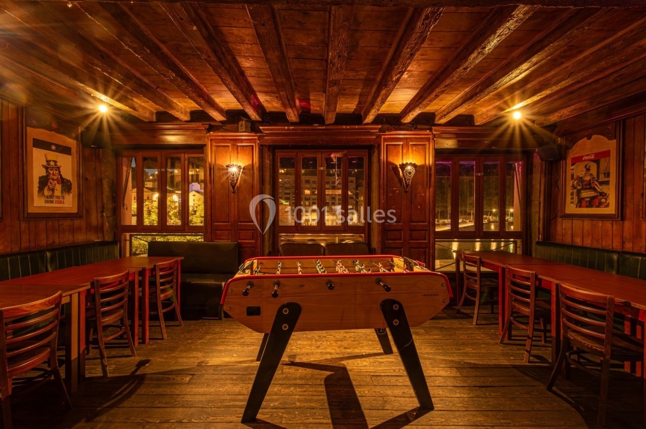 Salle en bois chaleureux avec un baby-foot au centre, entourée de tables et chaises, éclairage tamisé.