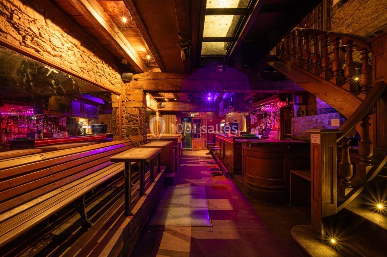 Intérieur d'un bar chaleureux avec éclairage tamisé, comptoir en bois, bancs alignés et escalier en colimaçon.