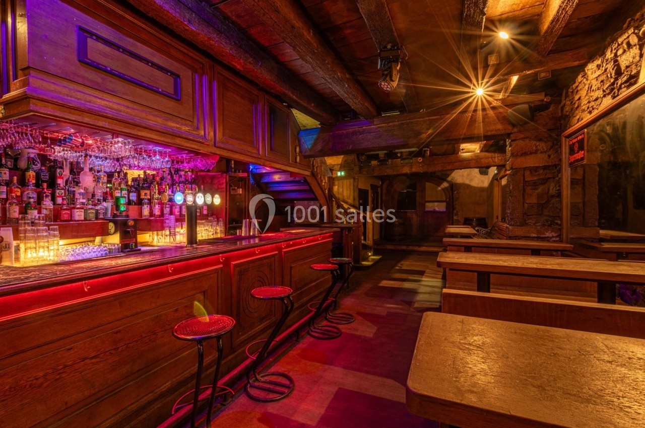 Bar en bois avec comptoir éclairé, tabourets hauts, étagères de bouteilles et tables en bois dans une ambiance chaleureuse.