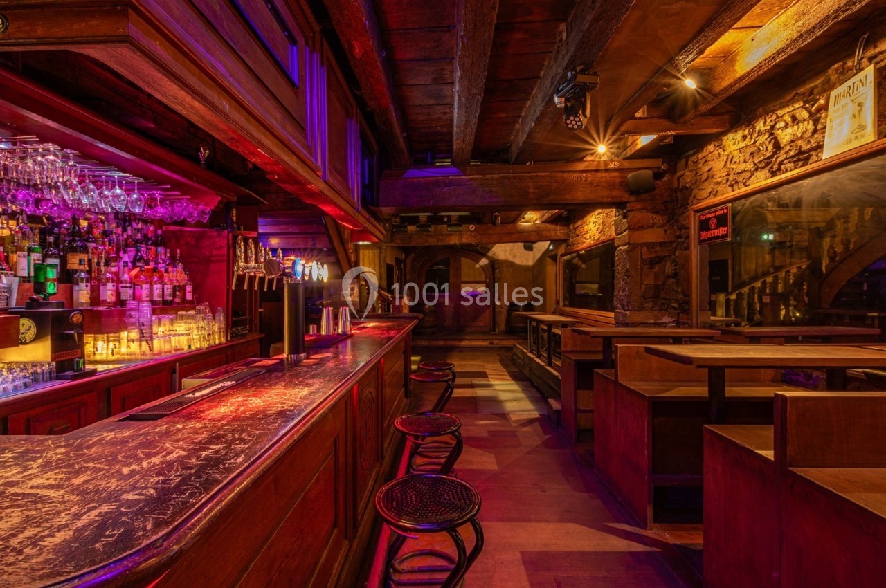 Intérieur d'un bar en bois avec un comptoir éclairé, des tabourets et des tables dans une ambiance chaleureuse.