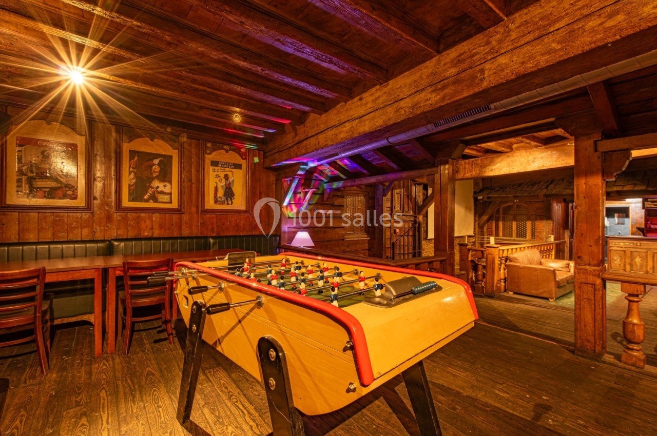 Baby-foot en bois dans une salle rustique avec poutres apparentes, éclairage chaleureux et décorations murales.