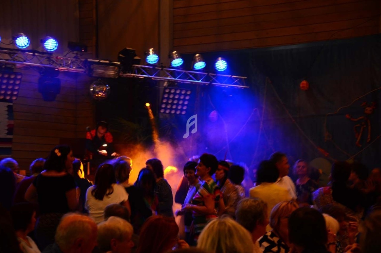 Groupe de personnes dansant dans une salle éclairée par des lumières colorées et un DJ en arrière-plan.