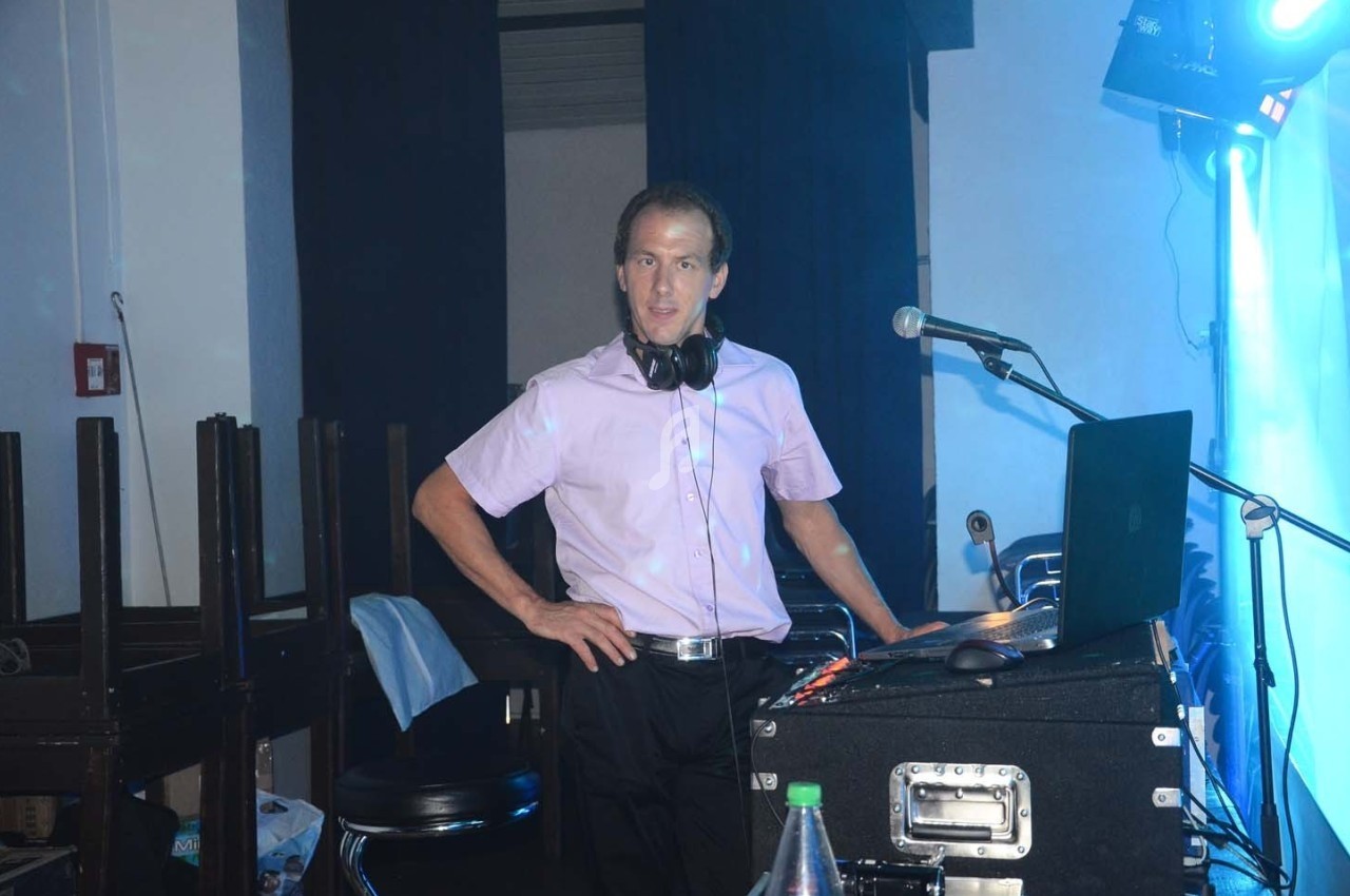 Homme debout derrière une table de mixage avec un ordinateur portable, portant un casque audio autour du cou.