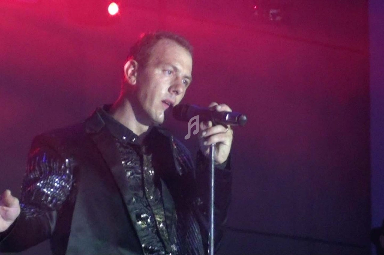 Un homme chante sur scène avec un micro, éclairé par une lumière rouge dans une ambiance sombre.