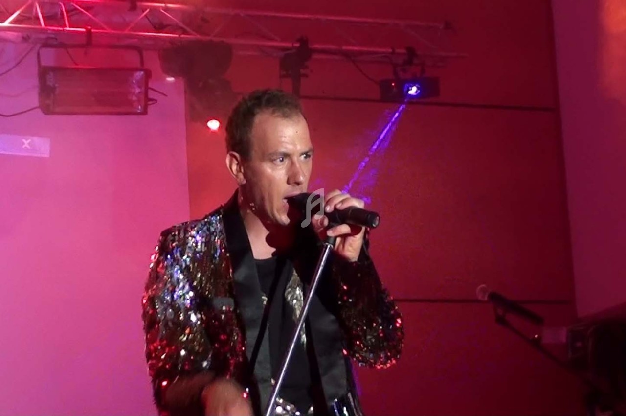 Homme chantant sur scène avec un micro, vêtu d'une veste à paillettes, éclairé par des lumières colorées.