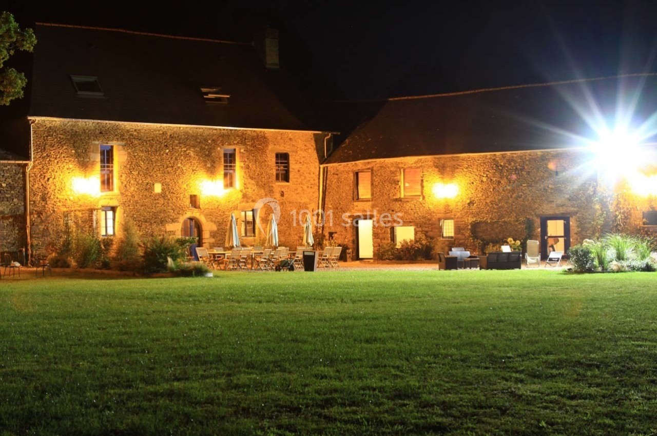 Bâtisse en pierre éclairée la nuit, entourée d'une pelouse et de mobilier de jardin disposé en terrasse.