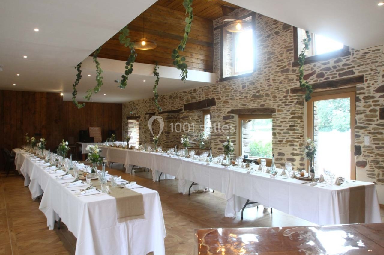 Salle de réception lumineuse avec murs en pierre, tables dressées en U et décoration végétale suspendue.