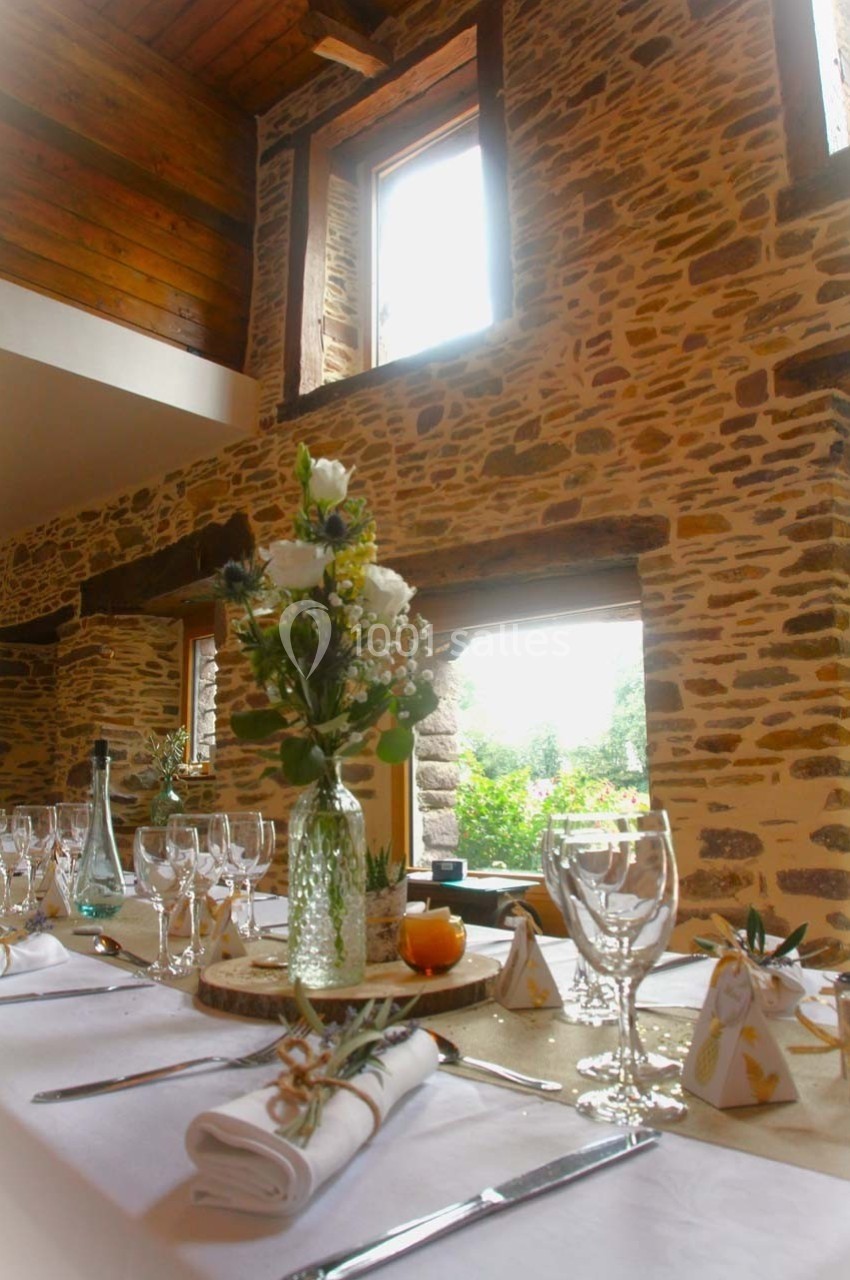 Table dressée avec élégance dans une salle en pierre apparente, décorée de fleurs et baignée de lumière naturelle.