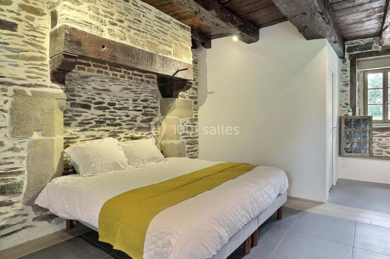 Lit double avec draps blancs et plaid jaune dans une chambre à murs en pierre apparente et poutres en bois au plafond.