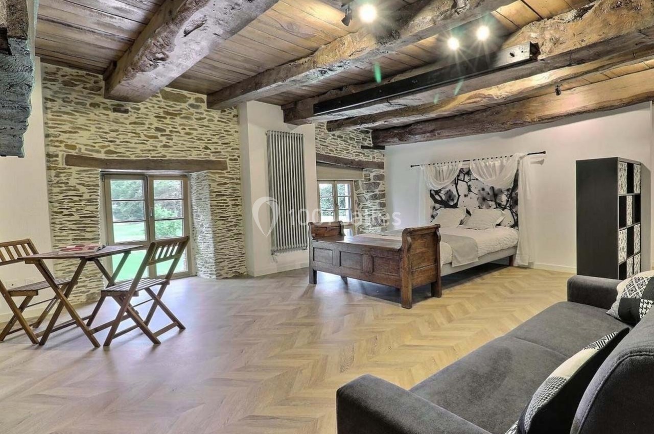 Grande pièce lumineuse avec murs en pierre, poutres apparentes, lit double, table en bois et canapé gris.