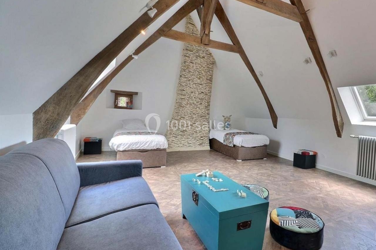 Chambre mansardée avec poutres apparentes, deux lits simples, canapé gris et coffre bleu sur un sol en parquet.