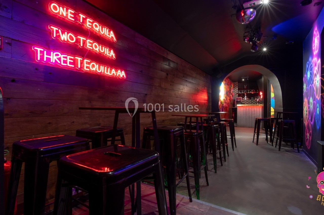 Salle de bar avec murs en bois, néons roses, tabourets hauts et comptoir éclairé au fond.