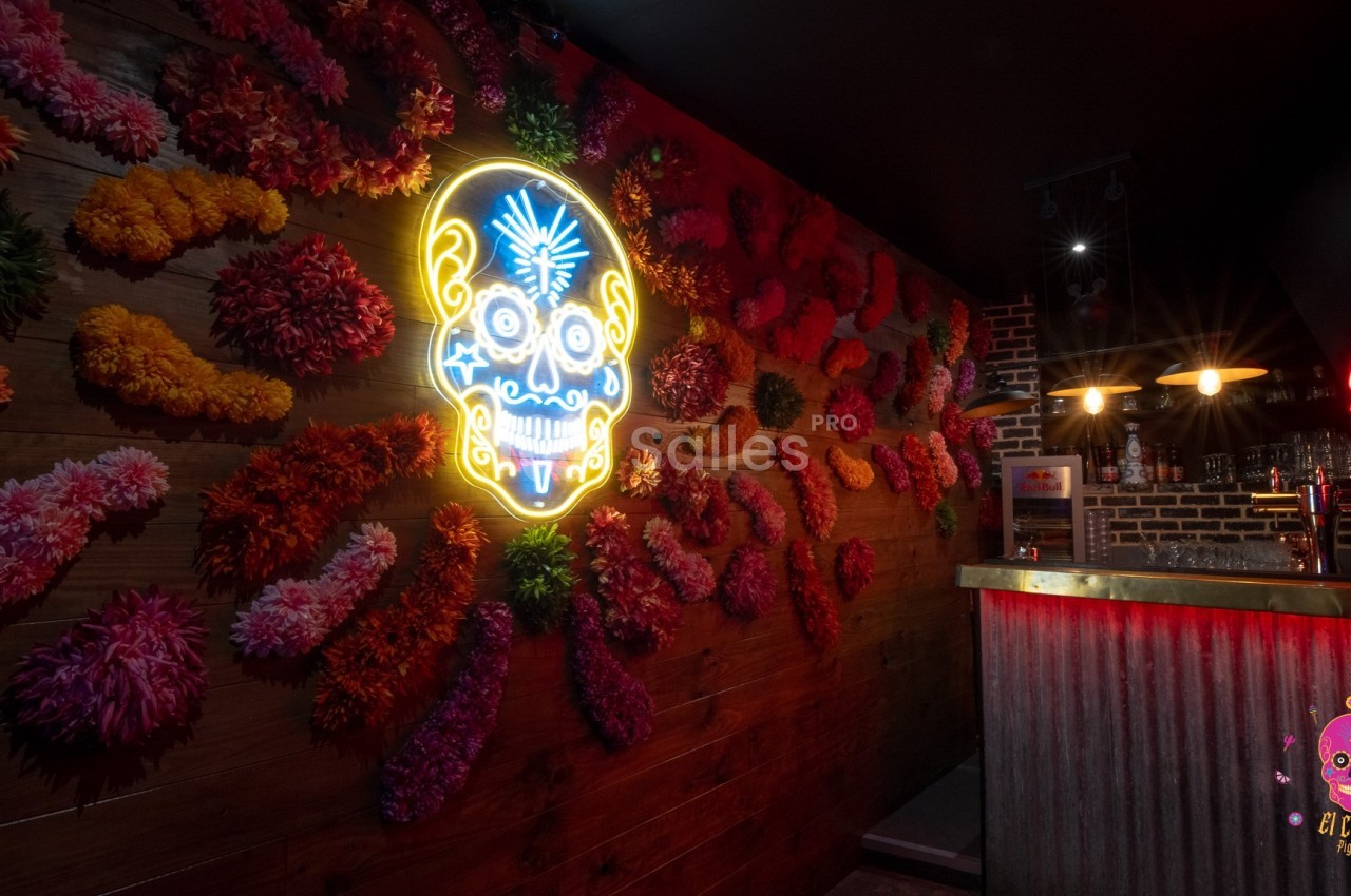 Mur décoré de fleurs colorées et d'une enseigne néon en forme de crâne, près d'un bar à l'éclairage tamisé.