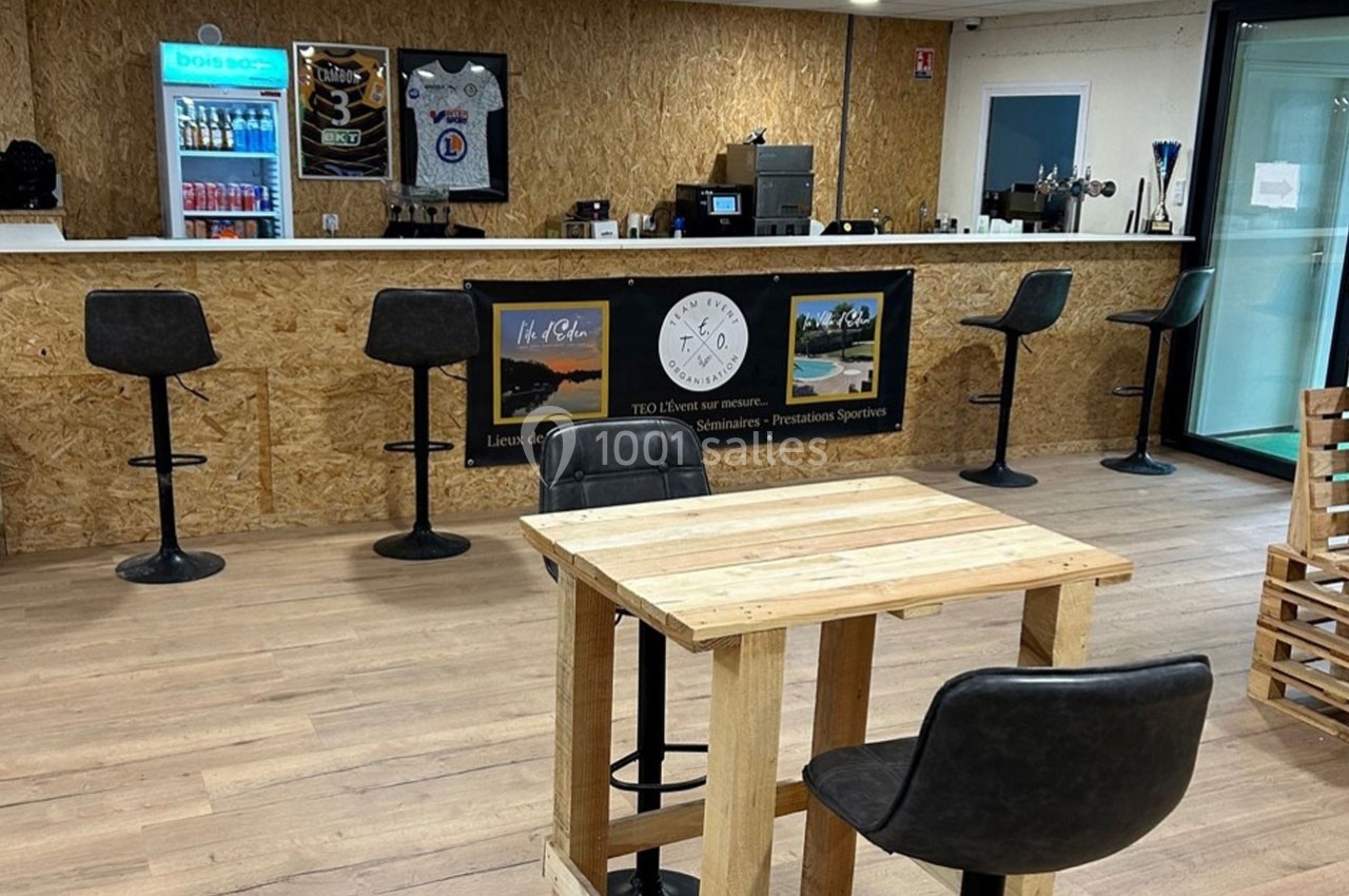 Bar avec comptoir en bois, tabourets hauts, table en bois clair au premier plan et décoration sportive en arrière-plan.