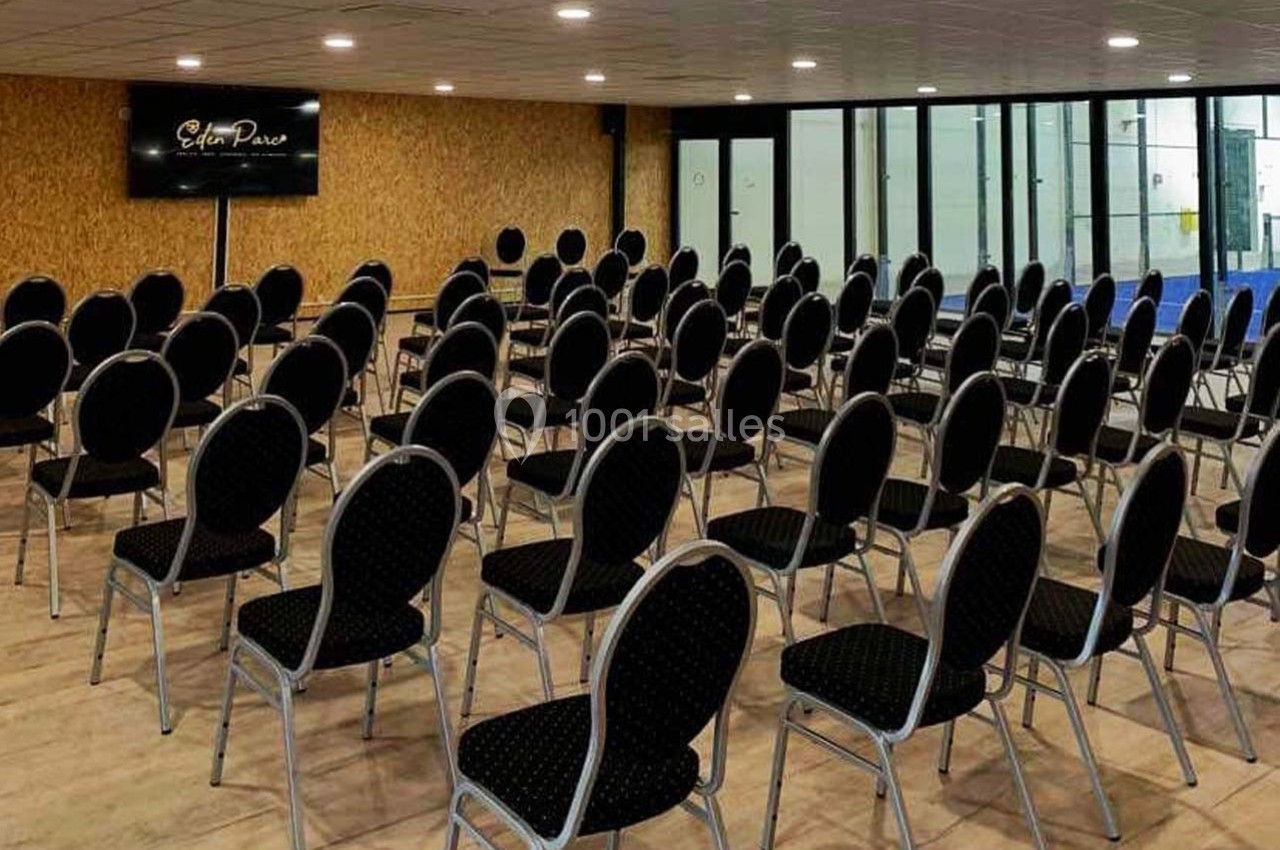 Salle de conférence avec des rangées de chaises noires alignées devant un écran mural, sol en bois clair.