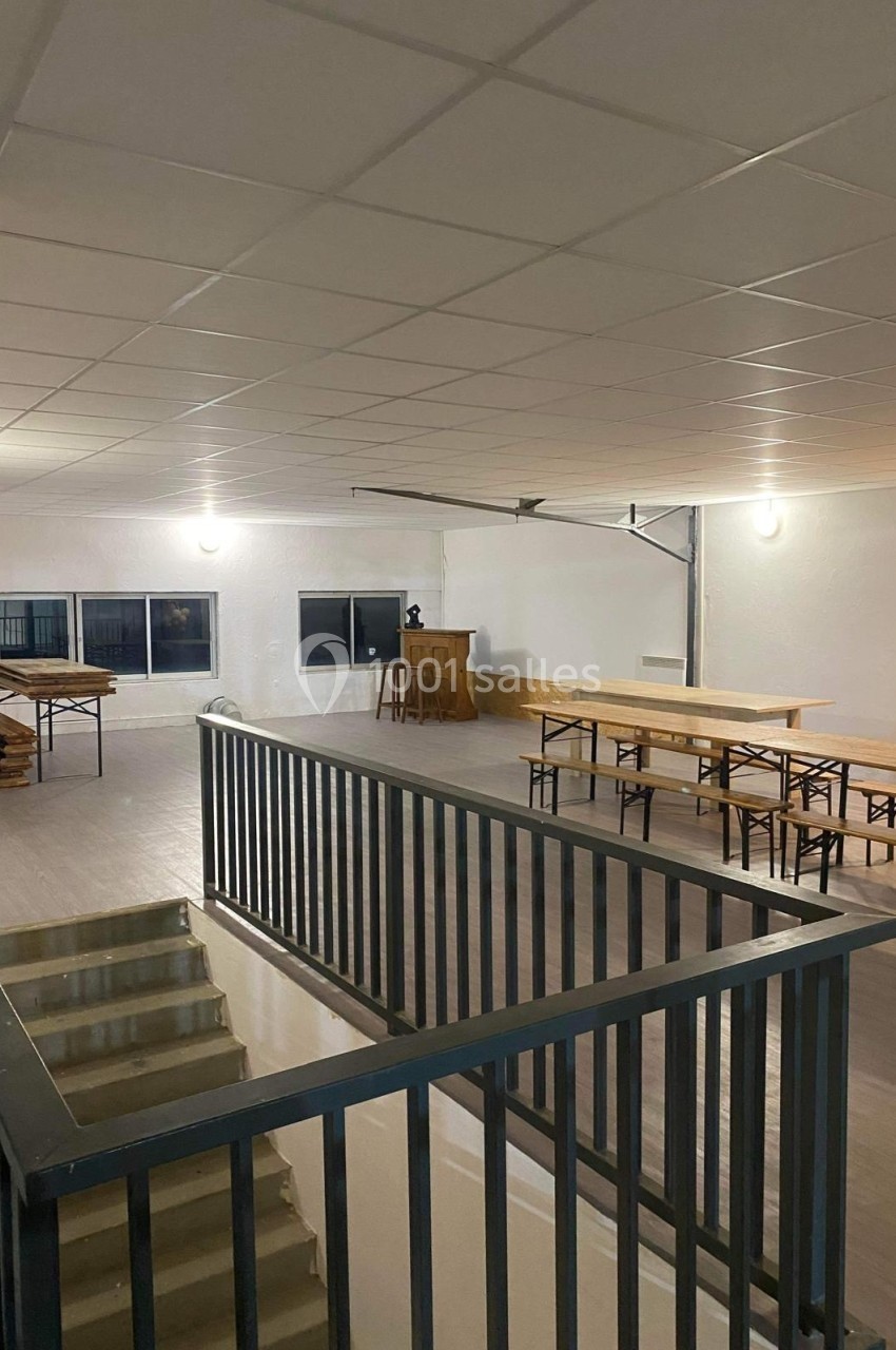 Salle spacieuse avec tables en bois, chaises pliantes et bar au fond, éclairée par des plafonniers et des fenêtres.