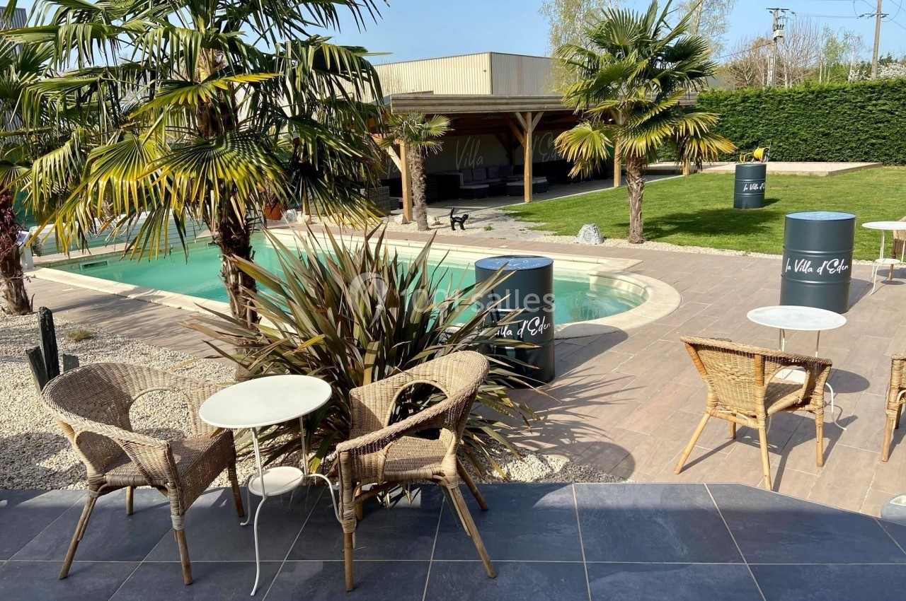 Terrasse avec tables et chaises en rotin, vue sur une piscine entourée de palmiers et d'un espace vert.