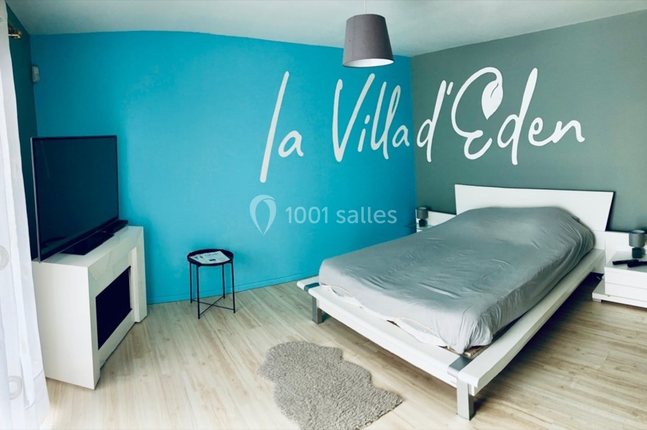 Chambre moderne avec un mur bleu, lit double, téléviseur, cheminée décorative et inscription ’La Villa d'Eden’.