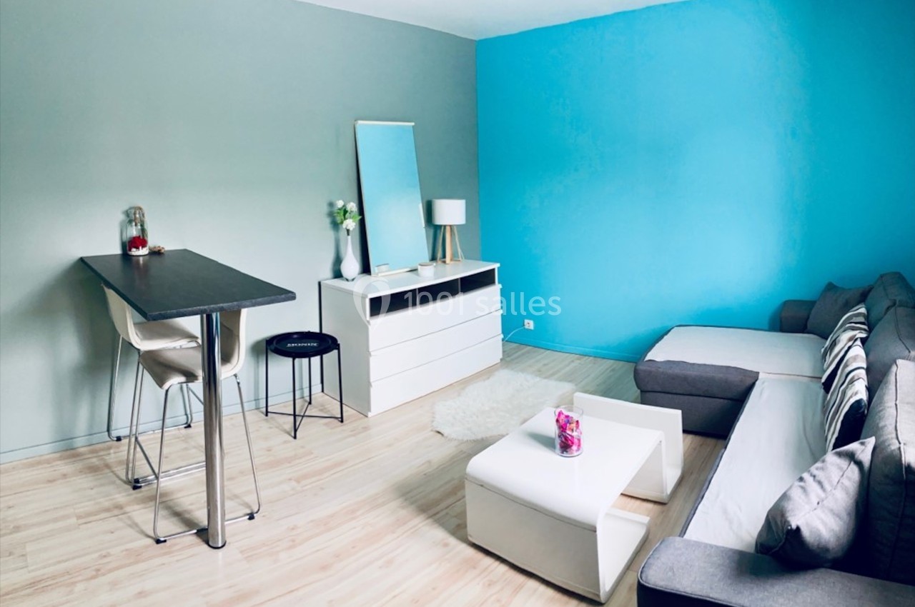 Salon moderne avec canapé gris, mur bleu, table haute avec tabourets, commode blanche et décoration minimaliste.