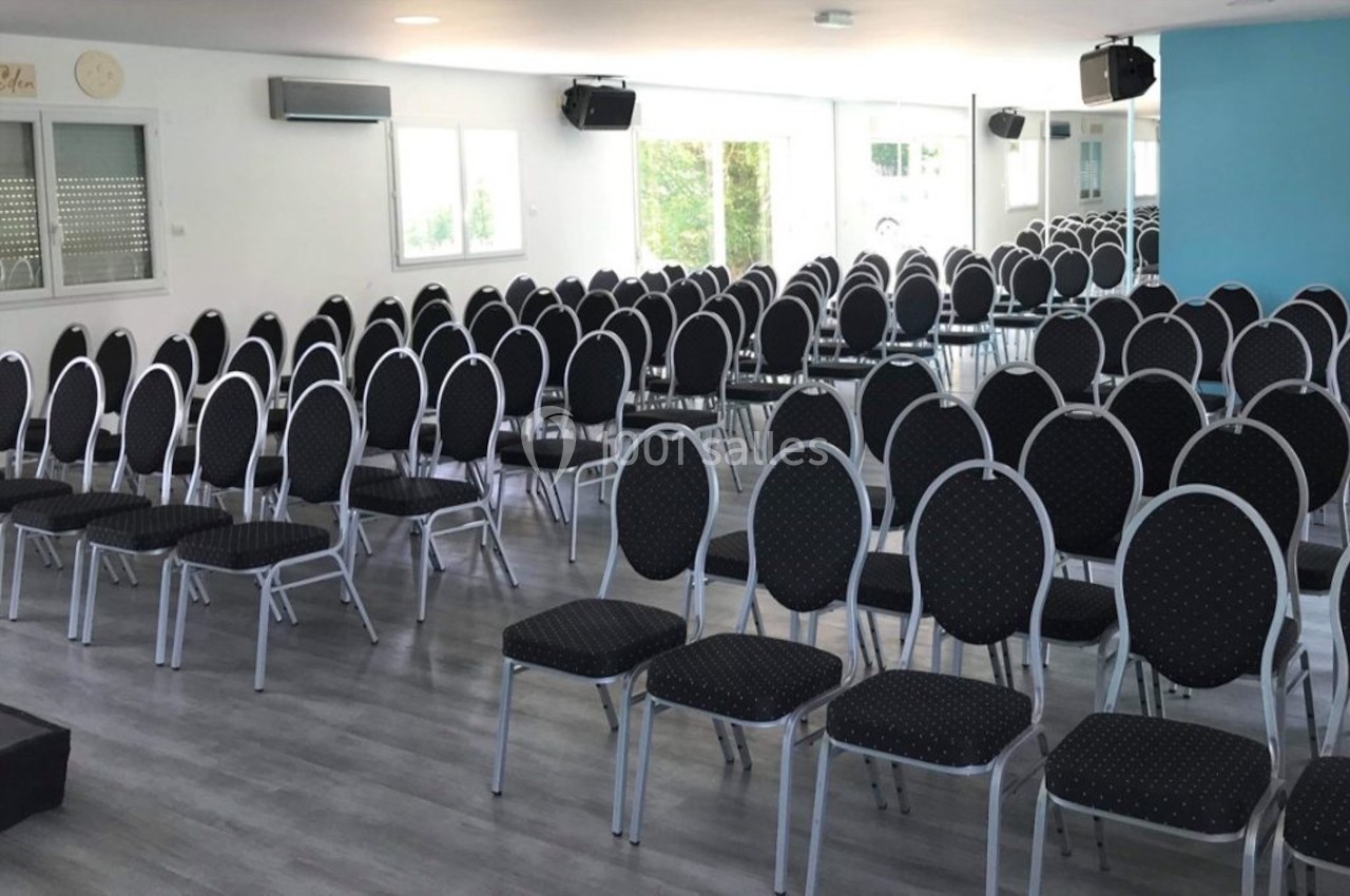 Salle lumineuse avec des rangées de chaises noires alignées sur un sol en parquet gris, prête pour un événement.
