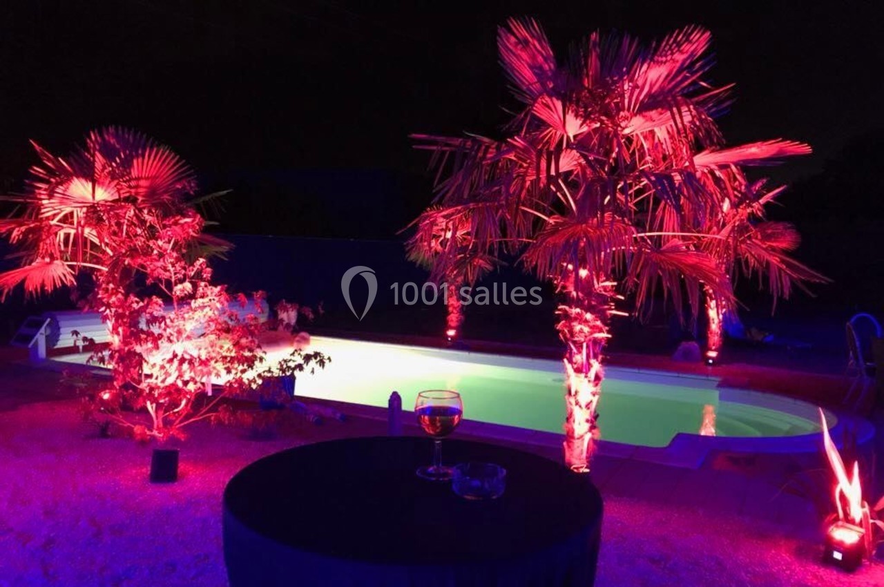 Piscine éclairée de nuit avec des lumières rouges et violettes, entourée de palmiers et d'une table avec un verre.