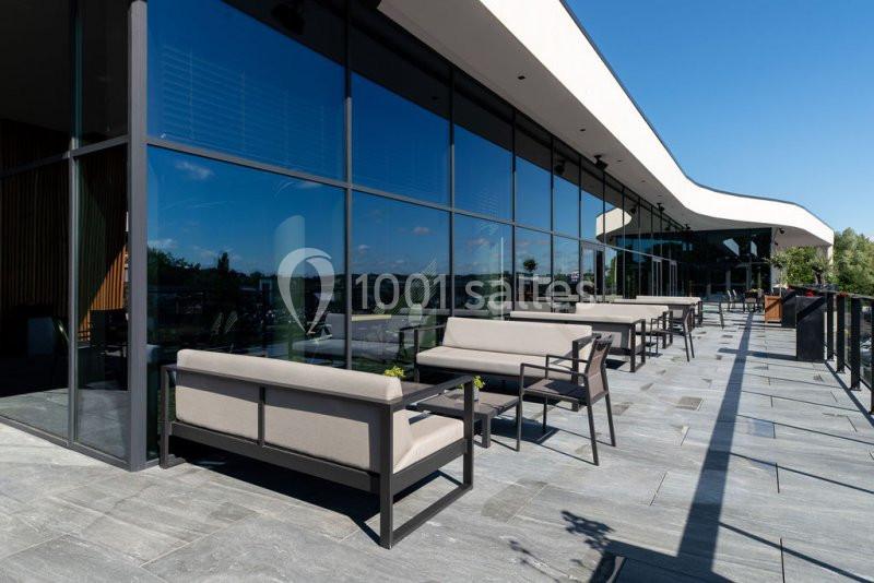 Location salle Nivelles (Brabant wallon) - Hôtel Nivelles Sud #8 Terrasse moderne avec mobilier en métal et coussins, bordée de grandes baies vitrées et offrant une vue dégagée.