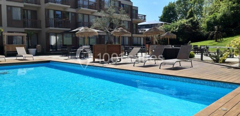 Location salle Nivelles (Brabant wallon) - Hôtel Nivelles Sud #18 Piscine extérieure entourée de transats, parasols en paille et bâtiments modernes, avec jardin en arrière-plan.