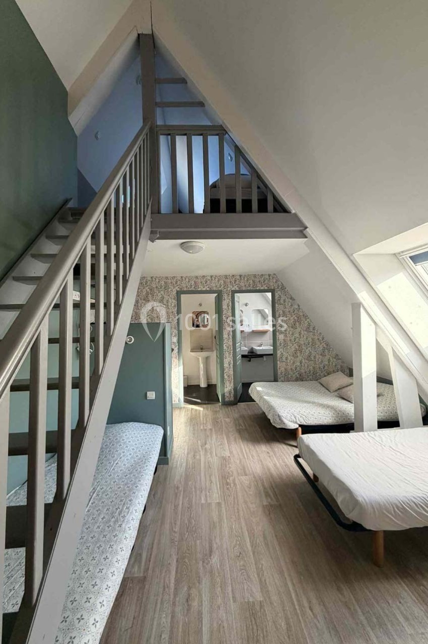 Chambre mansardée avec lits simples, parquet clair, murs verts et blancs, et escalier menant à une mezzanine.