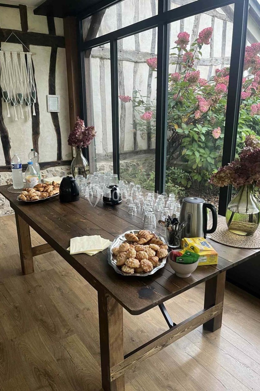 Table en bois avec biscuits, théière, tasses, bouteilles d'eau et fleurs, près d'une baie vitrée donnant sur un jardin.