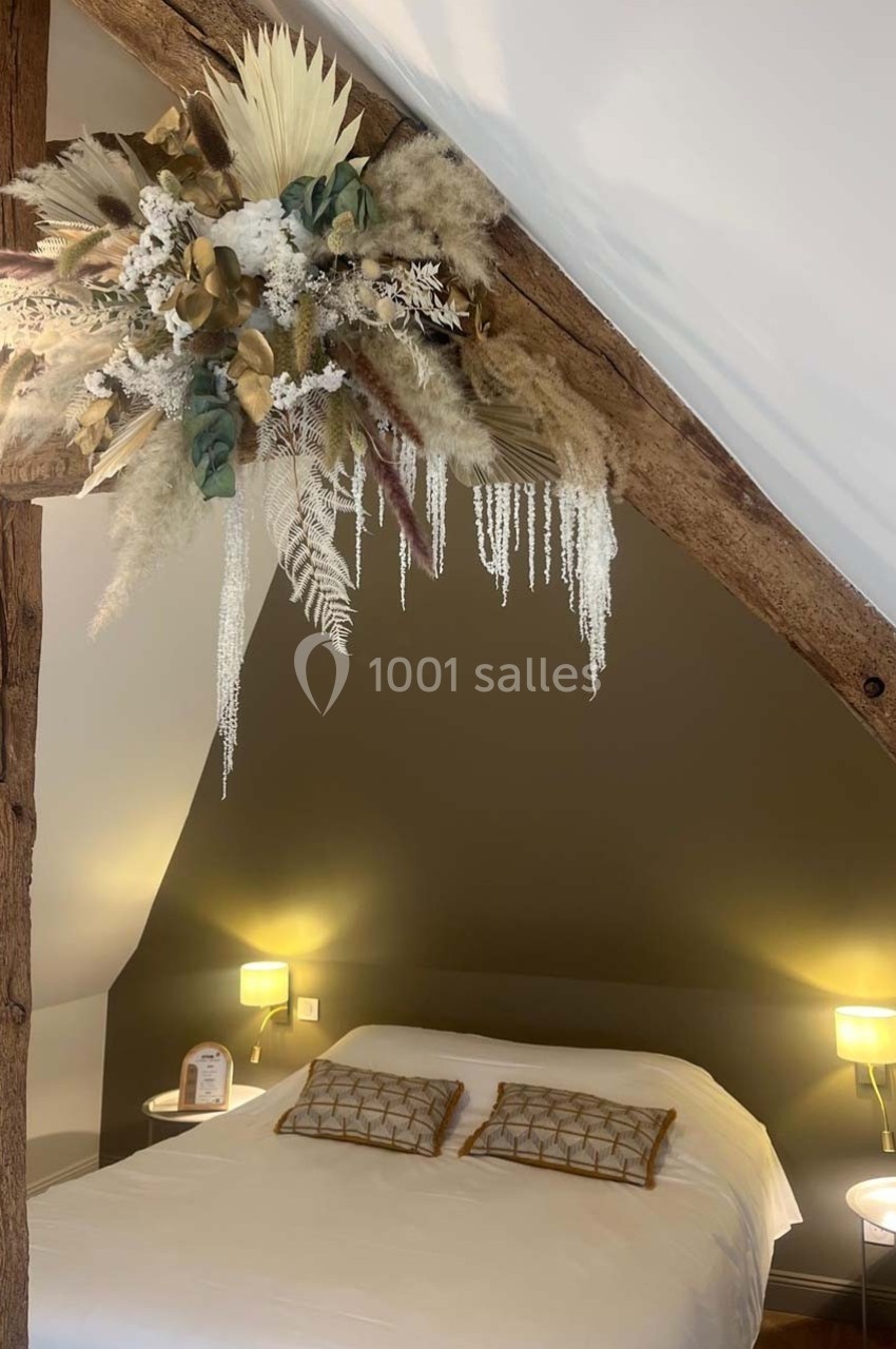 Lit double avec coussins décoratifs sous une poutre en bois ornée d'un bouquet de fleurs séchées, éclairage doux.