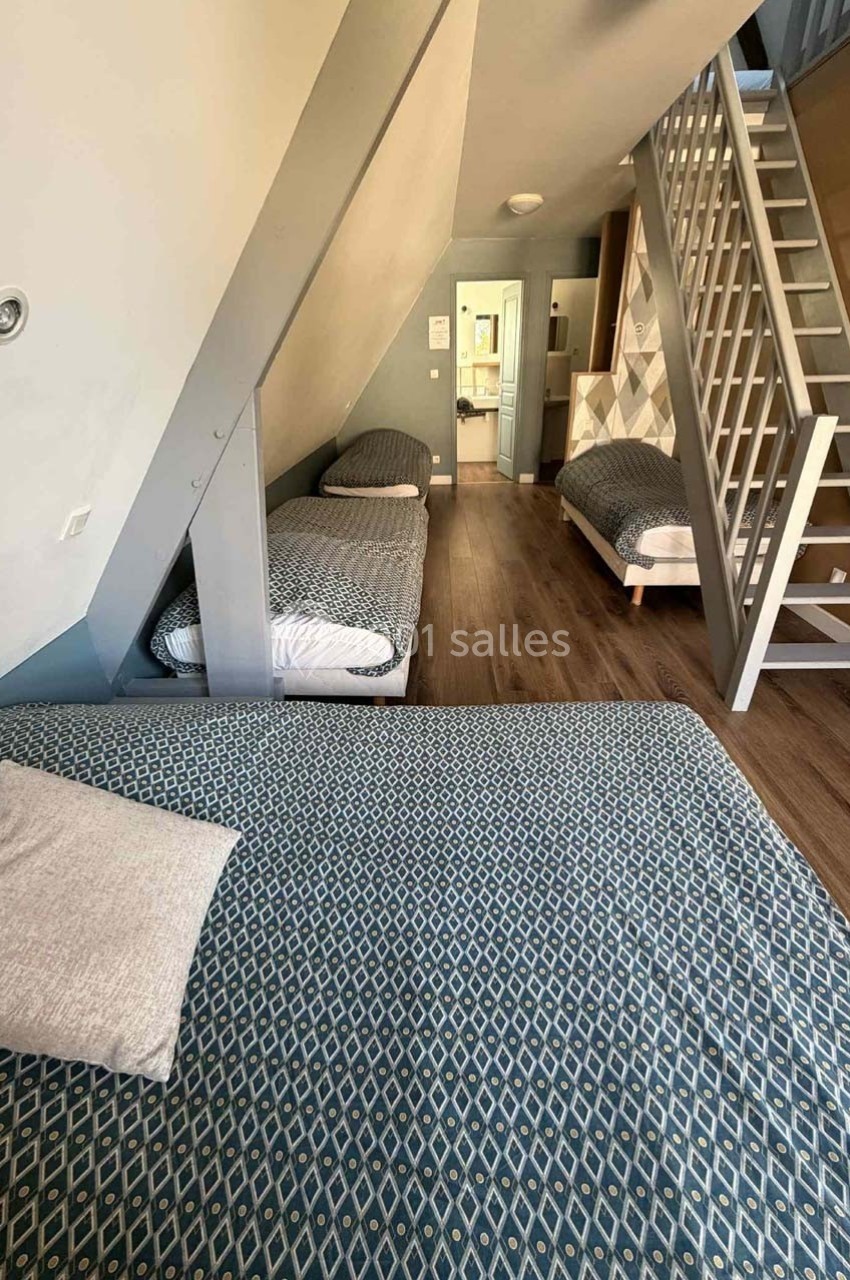 Chambre mansardée avec trois lits simples, parquet au sol, escalier menant à une mezzanine et lumière naturelle.