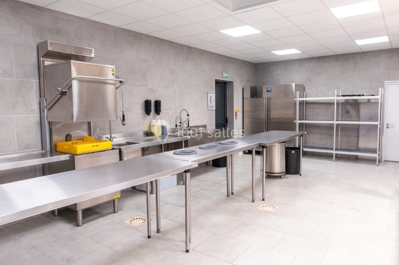 Cuisine professionnelle avec plans de travail en inox, lave-vaisselle industriel, étagères métalliques et réfrigérateur.