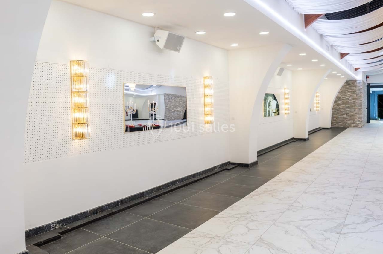 Couloir lumineux avec murs blancs, éclairages muraux modernes, sol en carrelage noir et blanc et plafond décoratif.